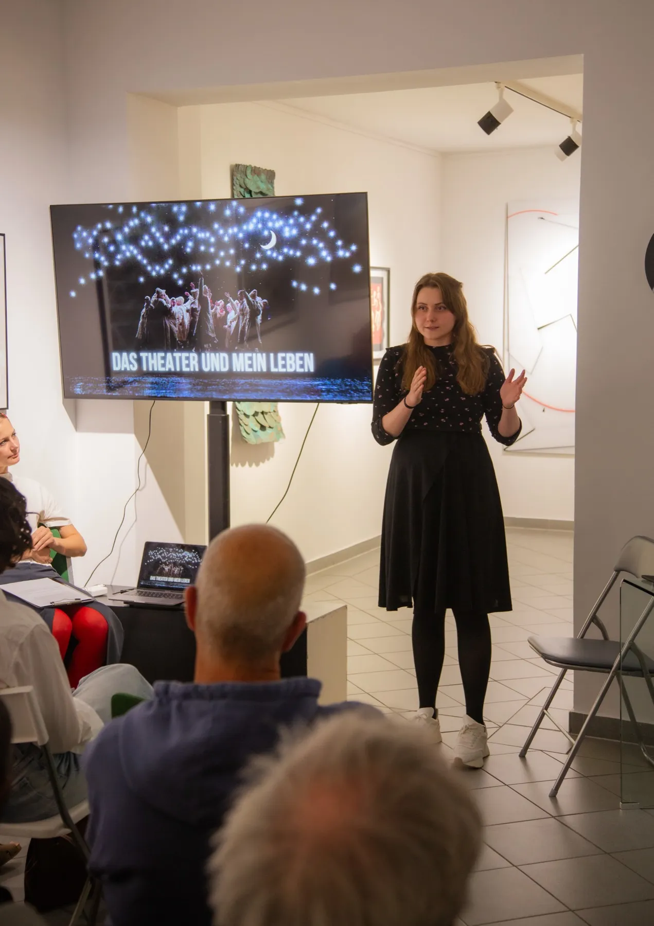 A speaker presents beside a screen reading “Theater ist mein Zuhause” while the audience listens.